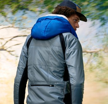 Junge Person, die sich abwendet, mit einer grauen Jacke und einer blauen Kapuze sowie einer Mütze, vor einem verschwommenen natürlichen Hintergrund aus Bäumen.