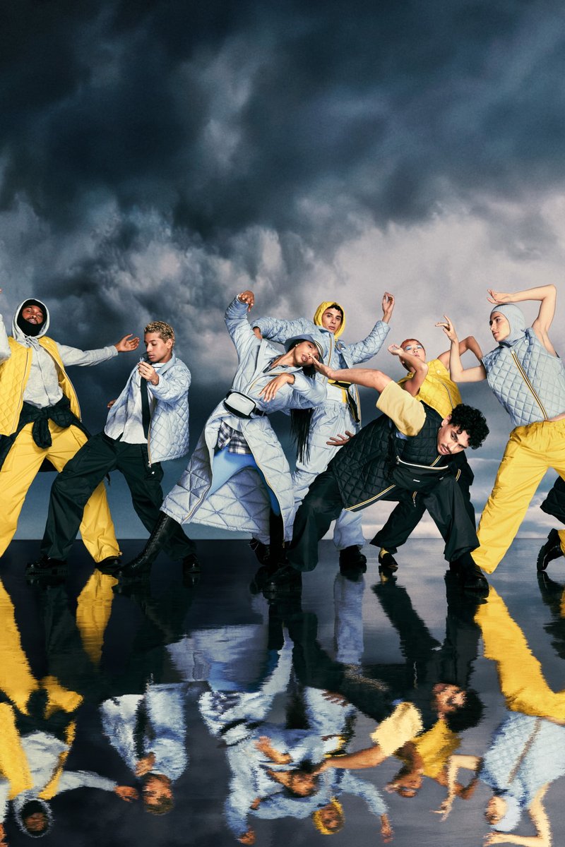 Nove persone vestite in modo elegante, con outfit vivaci, ballano espressivamente su uno sfondo drammatico e nuvoloso, mostrando movimento e stile.