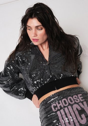 Frau mit langen dunklen Haaren, die eine schwarze Jacke mit Pailletten und eine Hose mit dem Schriftzug „CHOOSE JUICY“ in pinken Buchstaben trägt, schaut nach unten und zur Seite.