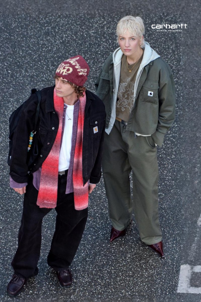 Two individuals stand on a patterned grey surface. One wears a black jacket, white shirt, red gradient scarf, and dark trousers. The other sports a green jacket and matching trousers.