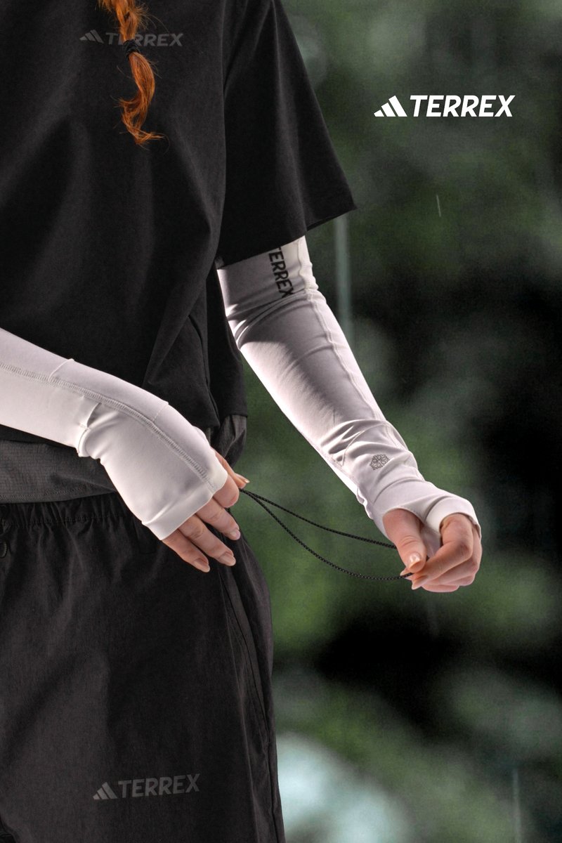 A person wearing a black shirt and white arm sleeves adjusts a drawstring on black pants, with a blurred green background.