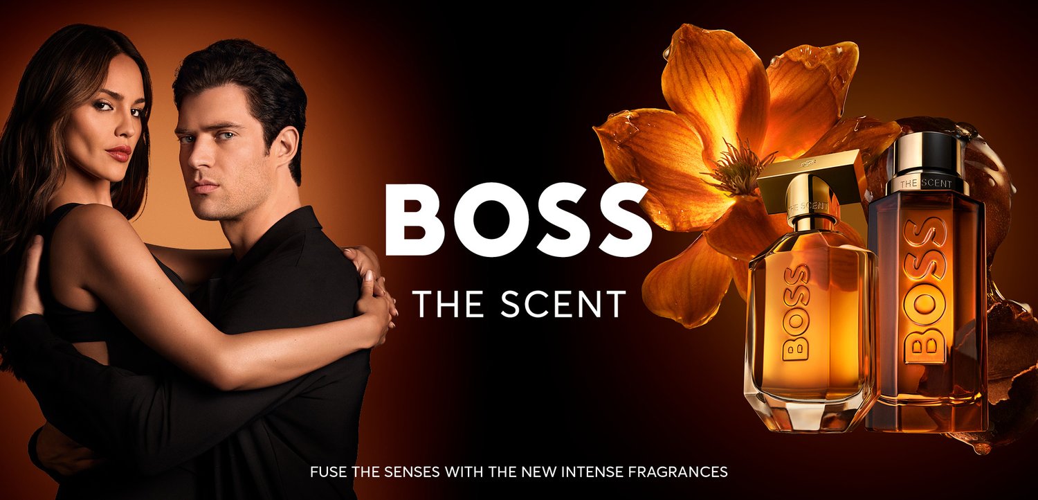 En mand og en kvinde omfavner hinanden foran en mørk orange baggrund med en stor blomst og to ravfarvede BOSS The Scent parfume flasker.