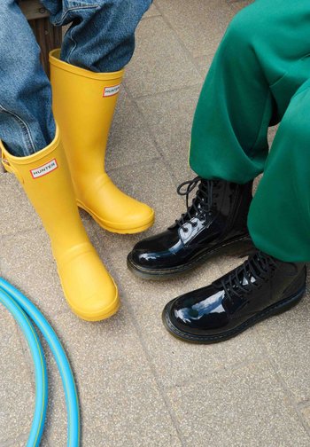 Deux personnes portant des bottes en caoutchouc jaunes et des bottines noires brillantes à lacets sont assises sur un sol carrelé avec des tuyaux d'arrosage bleus à proximité.