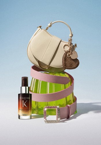 Beige handbag with heart keychains on a green glass container, pink leather belt with jeweled buckle, and Kérastase night serum bottle.
