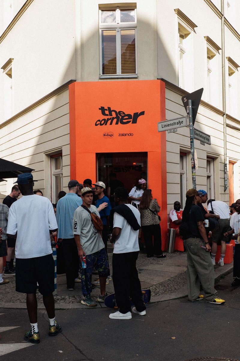 Udsigt til "hjørnet" butik med en orange facade. En mangfoldig gruppe mennesker er samlet udenfor og engageret i samtale. Vejskilte er synlige.