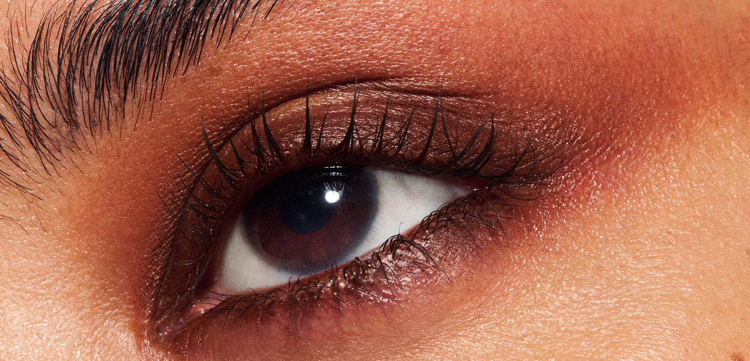 Gros plan sur un œil brun avec de longs cils noirs, entouré d'une peau lisse avec une teinte rougeâtre chaude et subtile et un léger éclat.