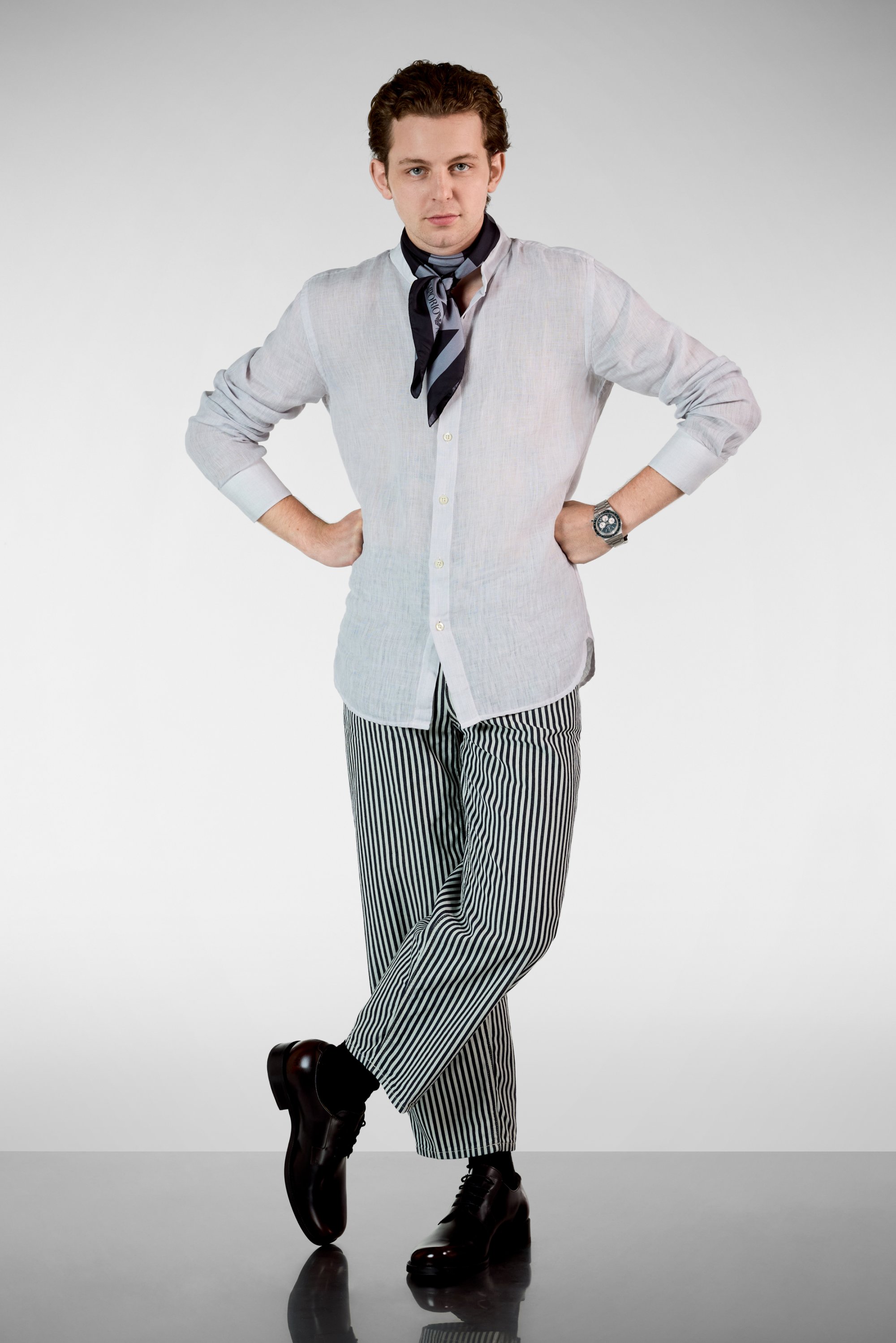 Camisa de lino gris claro con botones, pantalones rayados en blanco y negro, zapatos negros y una bufanda azul con estampado. Pose con las manos en las caderas.