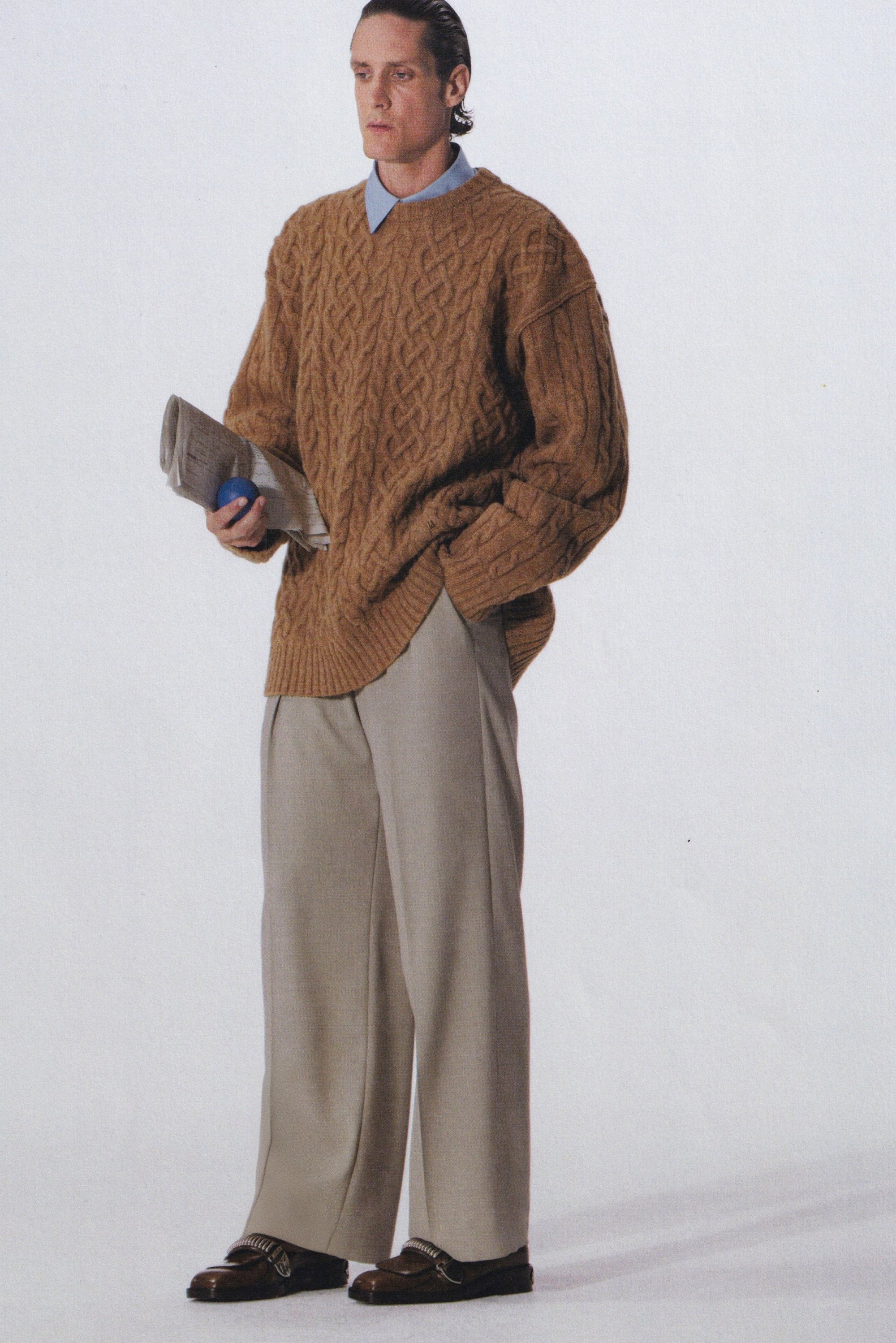 Overmaatse bruine kabelgebreide trui, lichtblauwe shirt eronder, beige wijde broek, met een krant en een blauwe bal in de hand. Bruine schoenen met accenten.
