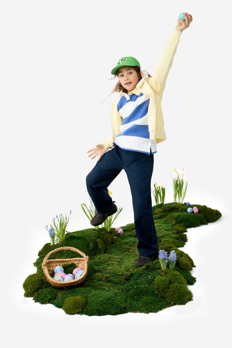 Child in green cap holding an Easter egg, standing on mossy ground with flowers and a basket of colorful eggs nearby.