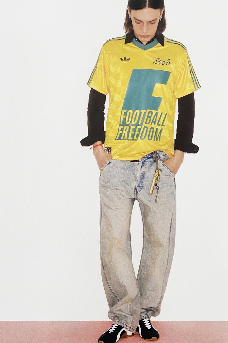 Personne portant un maillot jaune « Le football est liberté » par-dessus des manches longues noires, un jean ample délavé clair avec des breloques, et des baskets noires, debout les mains dans les poches.
