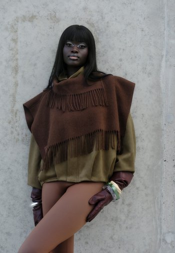 Brun poncho med frynse detaljer lagdelt over en grøn ruskindjakke. Modellen bærer bordeauxfarvede handsker og hudfarvede strømpebukser. Glatte, matte teksturer.