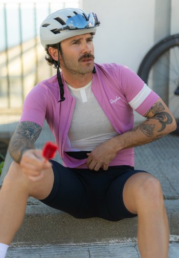 Cycliste moustachu en maillot rose et casque blanc assis sur le trottoir en train de manger une glace rouge, une roue de vélo visible derrière.
