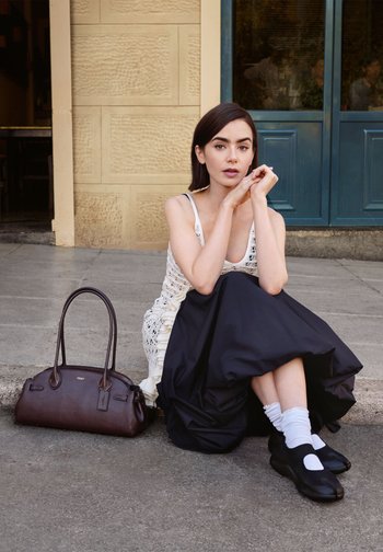 Young woman in white sleeveless top and black skirt sits on pavement beside brown leather handbag, in front of building with blue door.