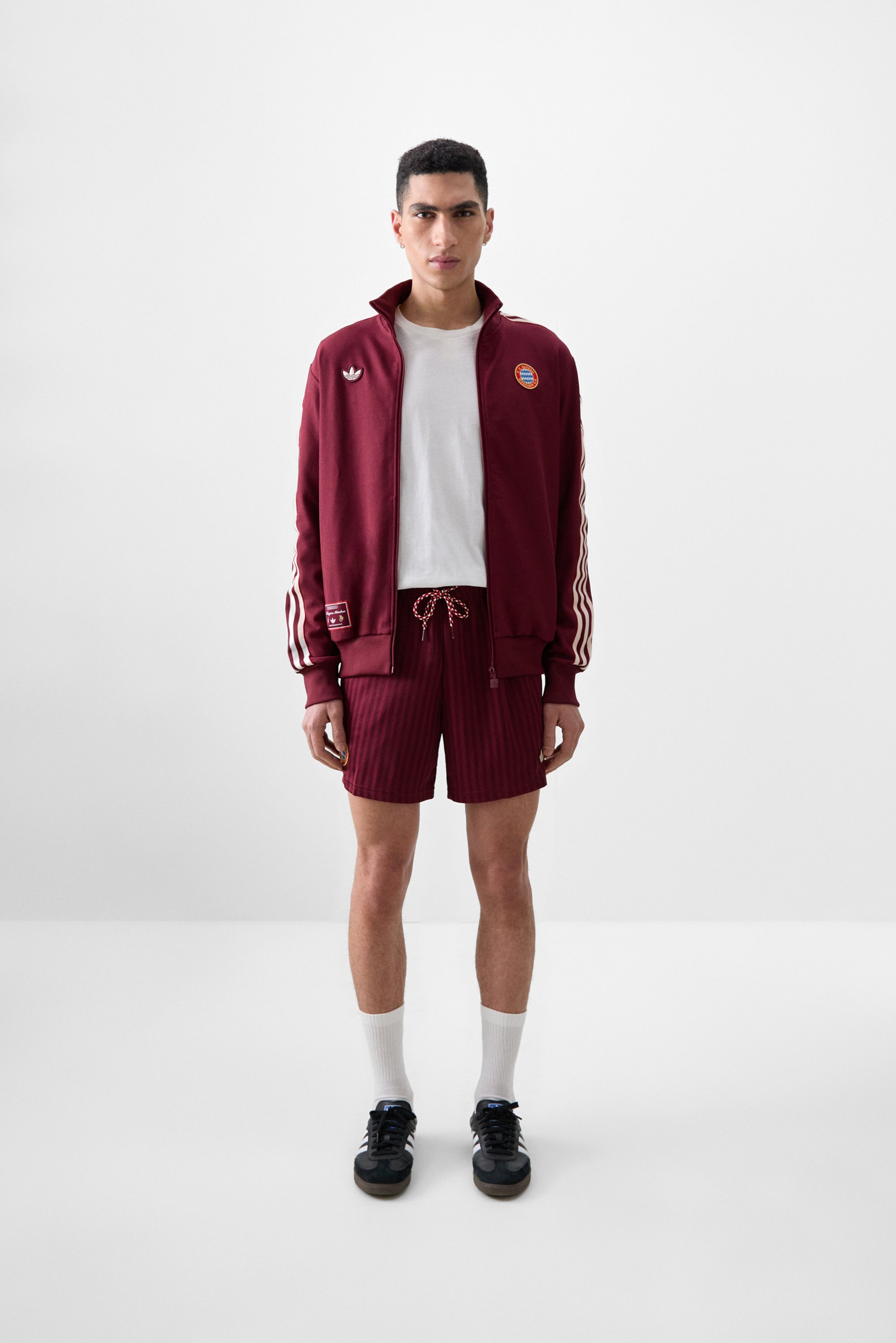 Jonge man staat zelfverzekerd, gekleed in een bordeauxrode trainingsjack, wit shirt, gestreepte shorts en zwarte sneakers, met een neutrale achtergrond.