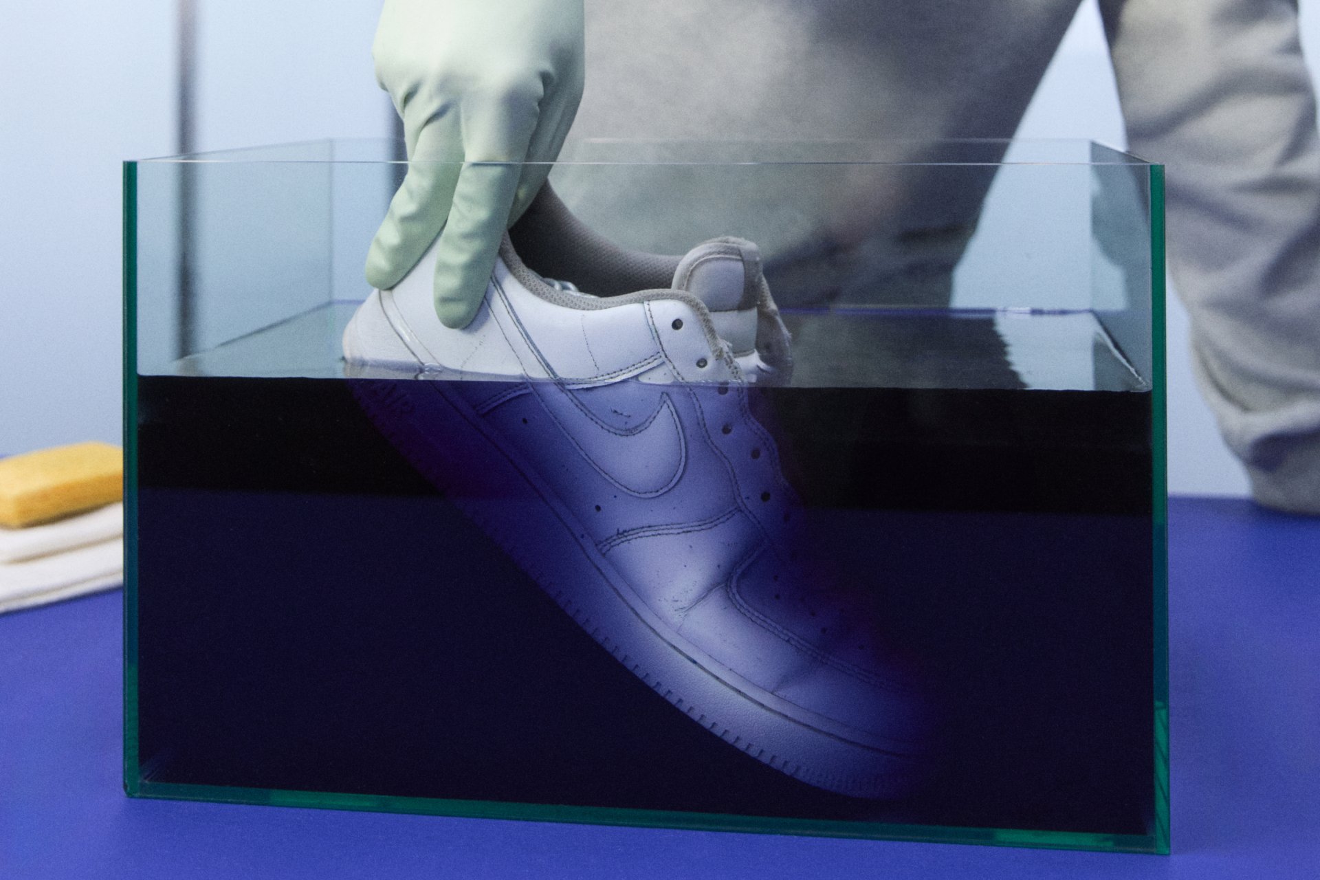Käsi yang memakai sarung tangan mencelupkan sepatu Nike putih ke dalam wadah kaca yang diisi dengan cairan gelap, yang diletakkan di atas permukaan biru.