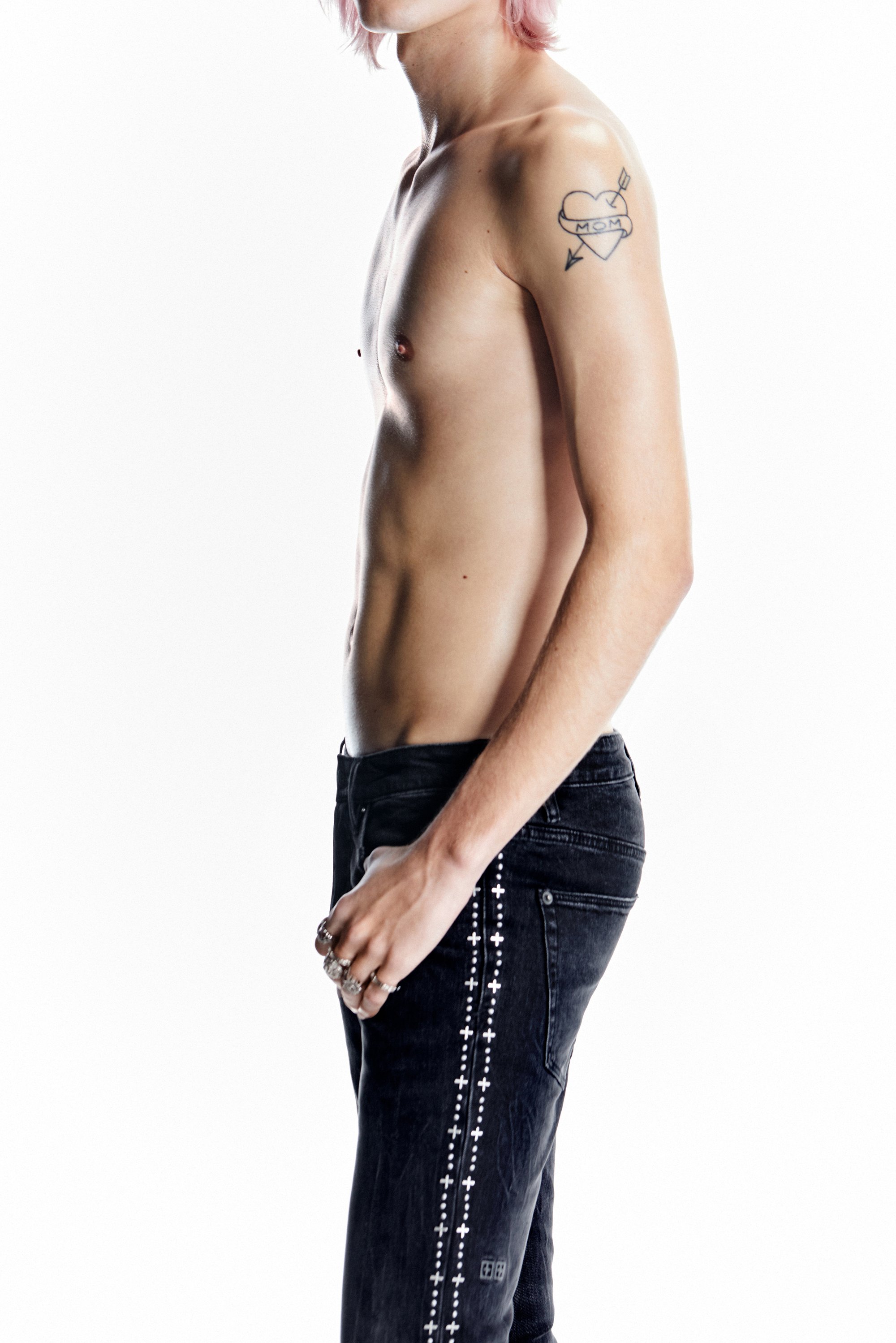A shirtless person with pink hair stands sideways, wearing black jeans with white decorative stitching and rings on their fingers.