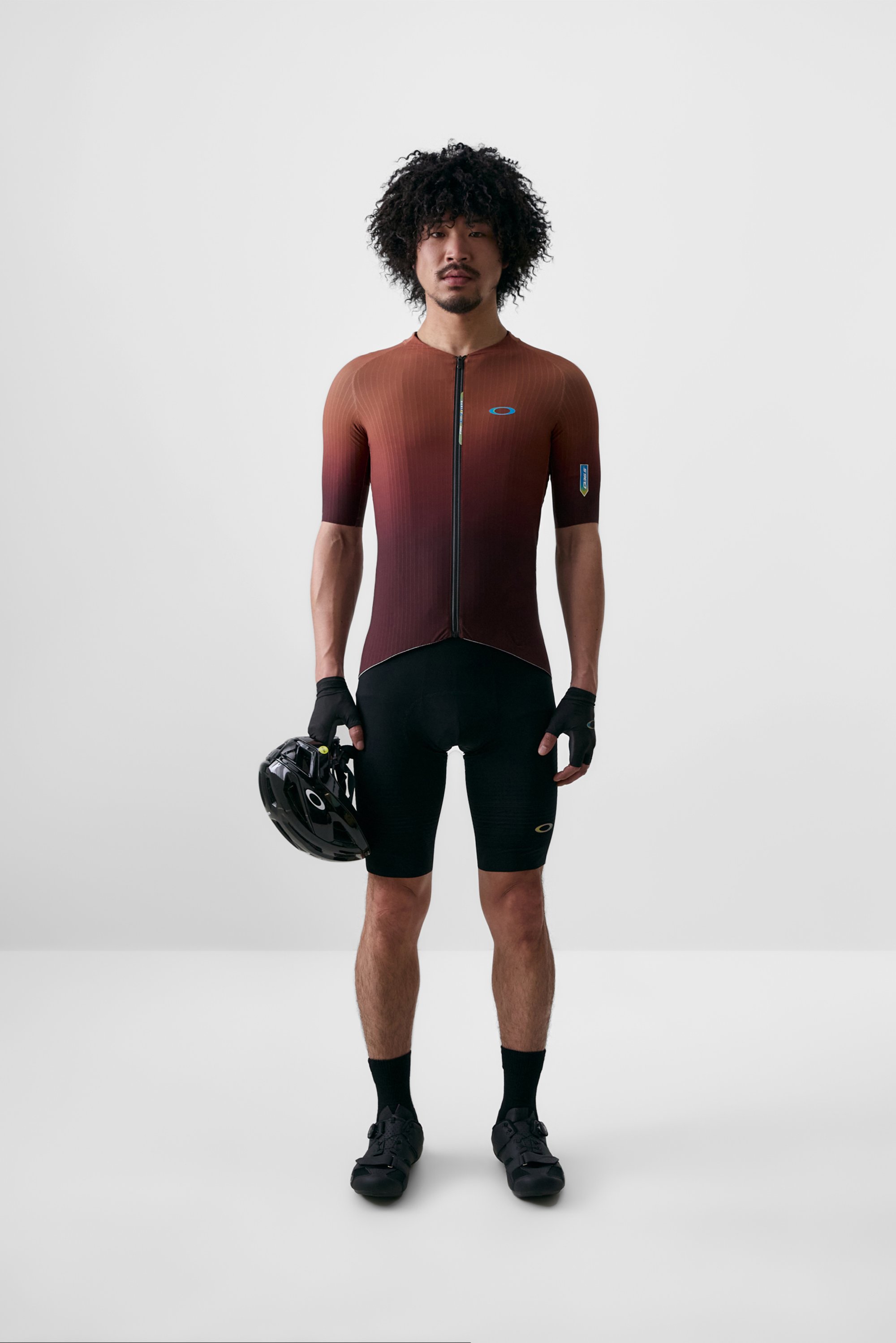 Un hombre con cabello rizado se presenta con confianza, vistiendo una camiseta de ciclismo con un degradado naranja y pantalones cortos, sosteniendo un casco negro con una mano.