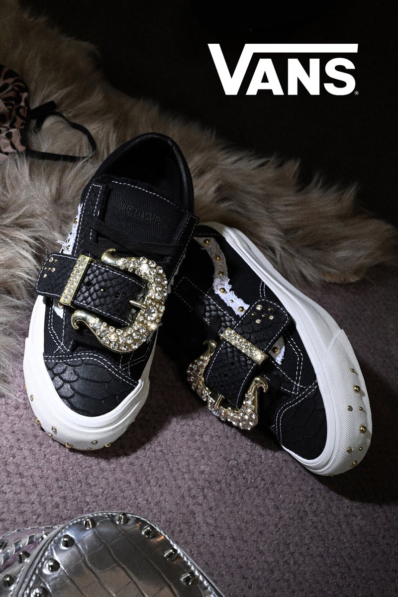 Schwarze Vans-Sneaker mit weißen Sohlen, großen goldenen Schmuckschnallen und dekorativen Nieten auf einem teppichbedeckten Boden mit einem Fellteppich in der Nähe.