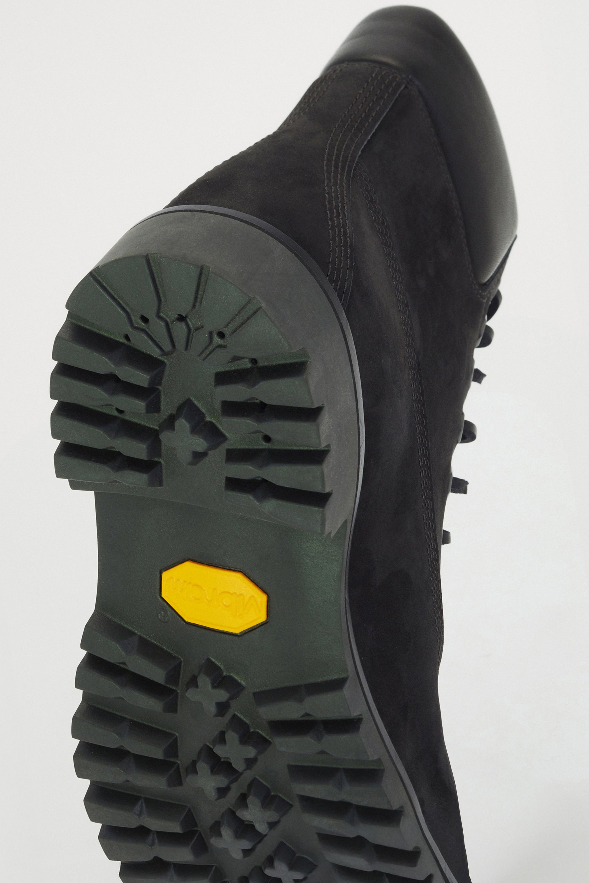 Black suede boot with a textured rubber sole. Features deep treads and a yellow Vibram logo. Smooth leather accents along the sides.
