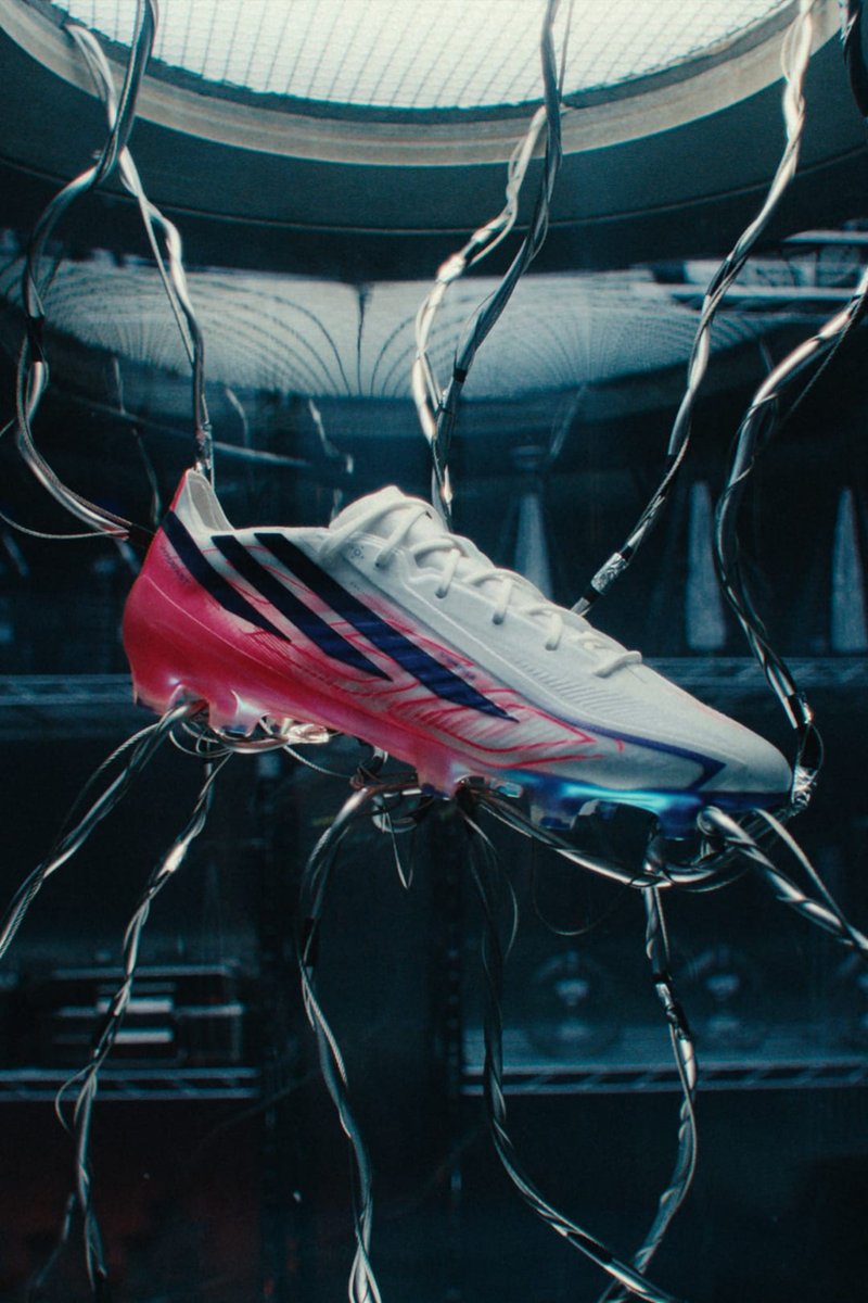 Weißer und roter Fußballschuh mit schwarzen Streifen, aufgehängt an verdrehten schwarzen Kabeln in einer dunklen Industrieumgebung.