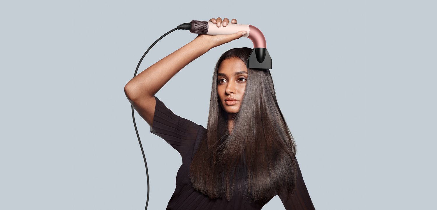 Fohn met een gebogen, roze handvat en zwarte mondstuk, vastgehouden om lang donker haar te steilen, tegen een lichtgrijze achtergrond.