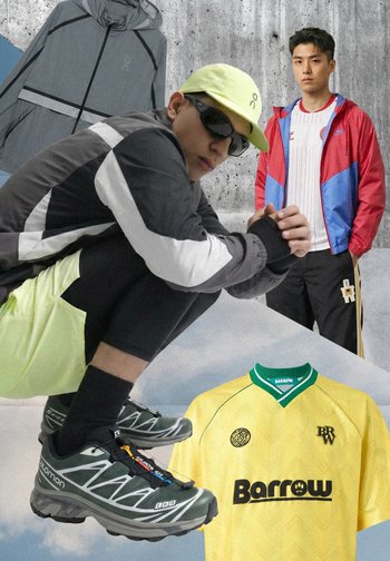 Zwei Männer tragen sportlich-legere Kleidung mit Jacken und Turnschuhen; gelbes Barrow-Trikot und graue Jacke vor Beton- und Himmelshintergrund.