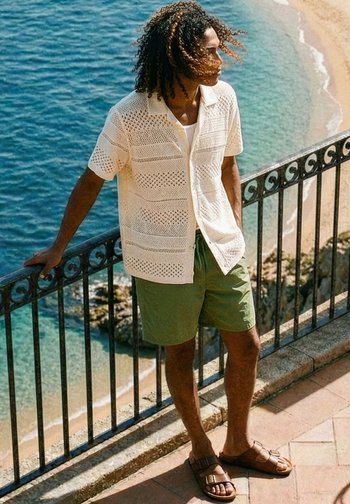 Homme aux cheveux bouclés portant un t-shirt blanc en maille à manches courtes, un short vert et des sandales marron, appuyé sur une rambarde près d’une plage et de l’océan.