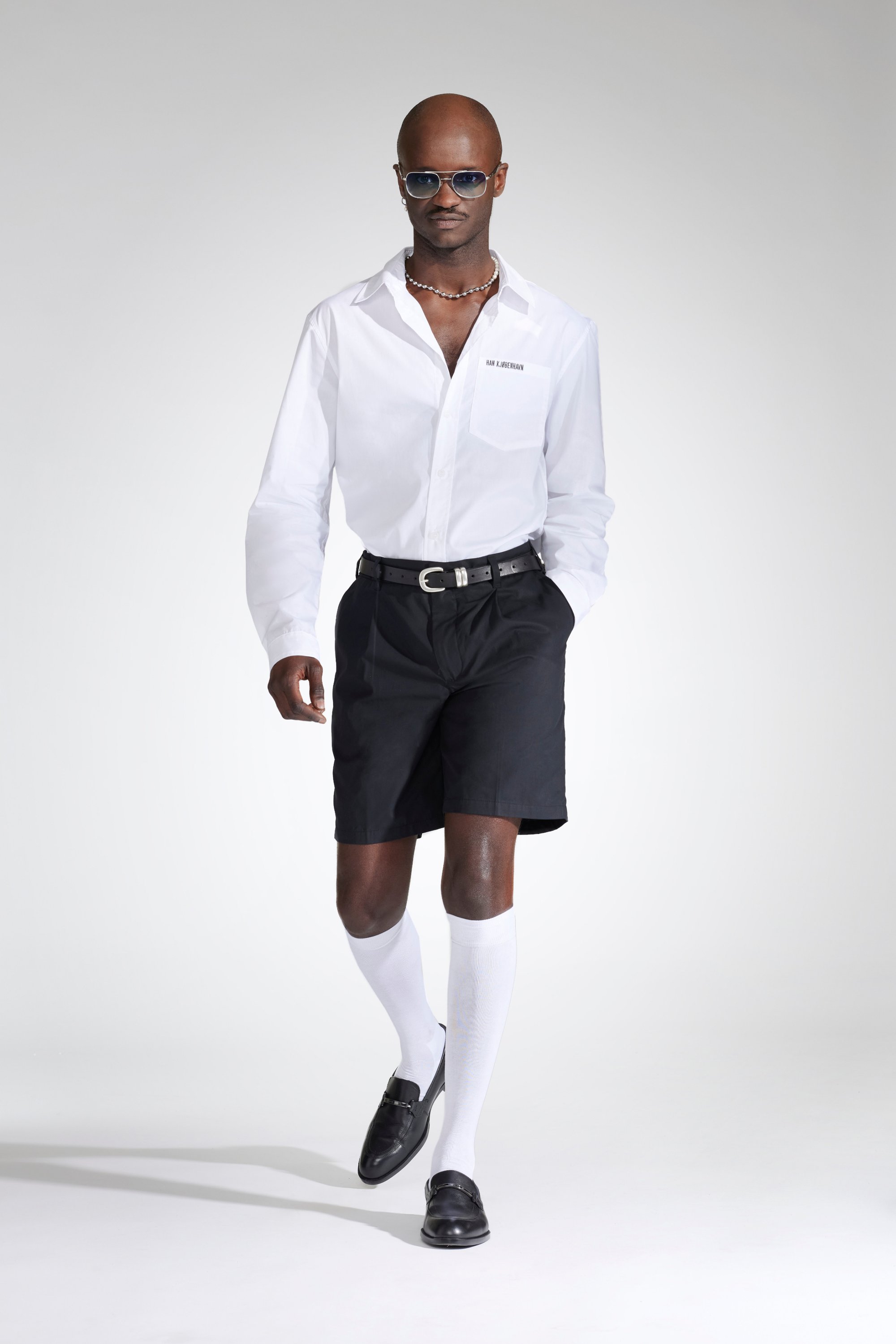 A man walks confidently in a white shirt, black shorts, knee-high white socks, and sunglasses, accessorised with a necklace.