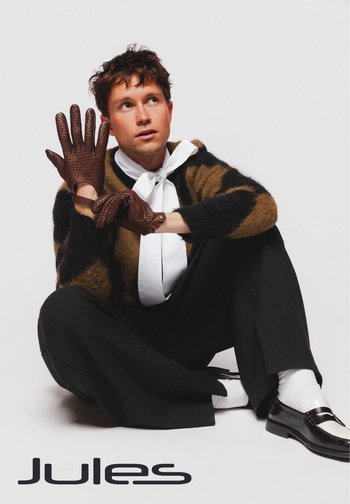 Gants en cuir marron à motif avec des doigts allongés, portés par une personne dans un pull rayé noir et marron, un pantalon noir et une chemise blanche.