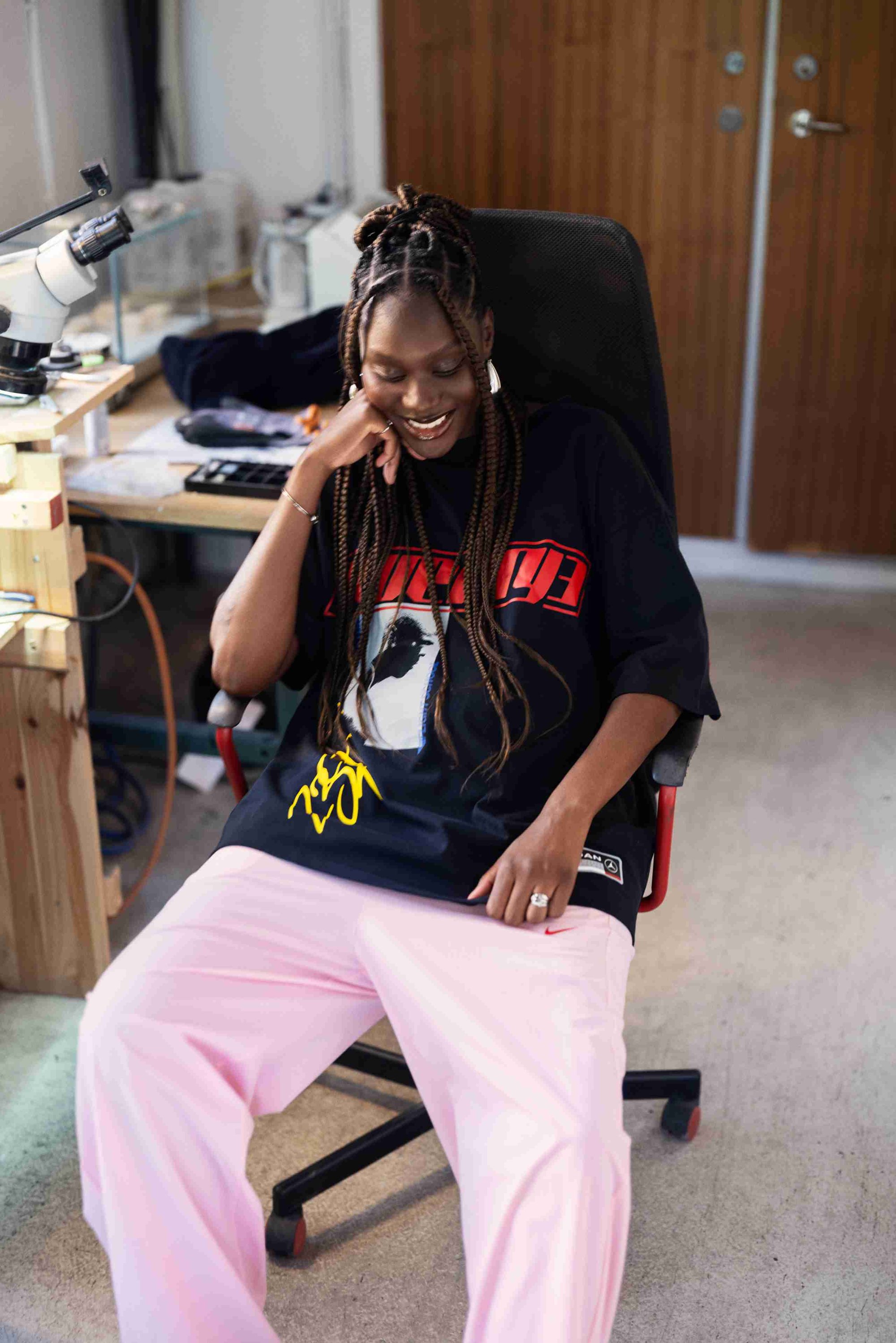 Zwart graphic t-shirt met rode tekst en een kleurrijke print, gecombineerd met losse roze broek. De setting bevat houten meubels en een microscoop.