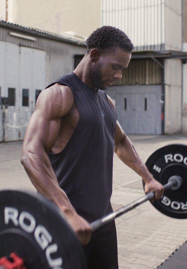 Un homme musclé portant un débardeur soulève une barre avec des poids en extérieur, concentré sur son entraînement avec une arrière-plan d'entrepôt.