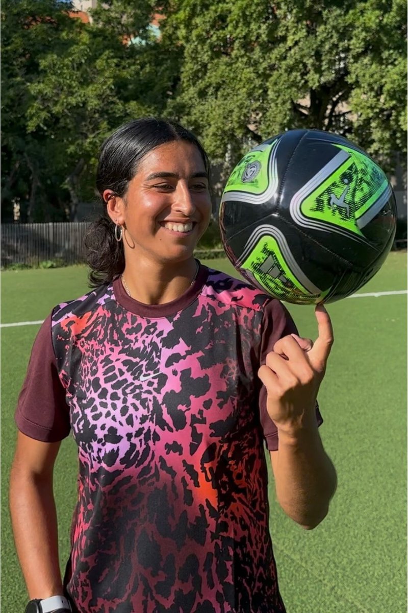 Woman smiling while balancing a black and green soccer ball spinning on her right index finger on a sunny outdoor soccer field.