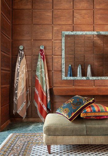 Δωμάτιο με ξύλινη επένδυση τοίχων, δύο πετσέτες με σχέδια κρεμασμένες, καθρέφτης με πλαίσιο από πράσινο μάρμαρο, μπεζ πάγκος και πολύχρωμα διακοσμητικά μαξιλάρια σε χαλί με σχέδια.