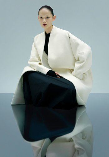 Übergroßer weißer Mantel aus glattem Stoff über einem schwarzen Kleid. Modell sitzt mit zurückgegelten Haaren und spiegelt sich auf einer glänzenden Fläche.
