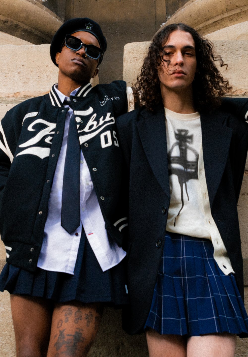 Zwei Personen stehen eng beieinander. Die eine trägt eine schwarze Collegejacke, ein weißes Hemd, eine blaue Krawatte und einen schmeichelhaften rock. Die andere trägt einen schwarzen Blazer über einem bedruckten T-Shirt und einem karierten Rock.