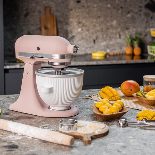 Jasnoróżowy mikser stojący ze stalową misą, o teksturowanej powierzchni i płaskiej podstawie, otoczony pokrojonymi owocami i przyborami do pieczenia na blacie.