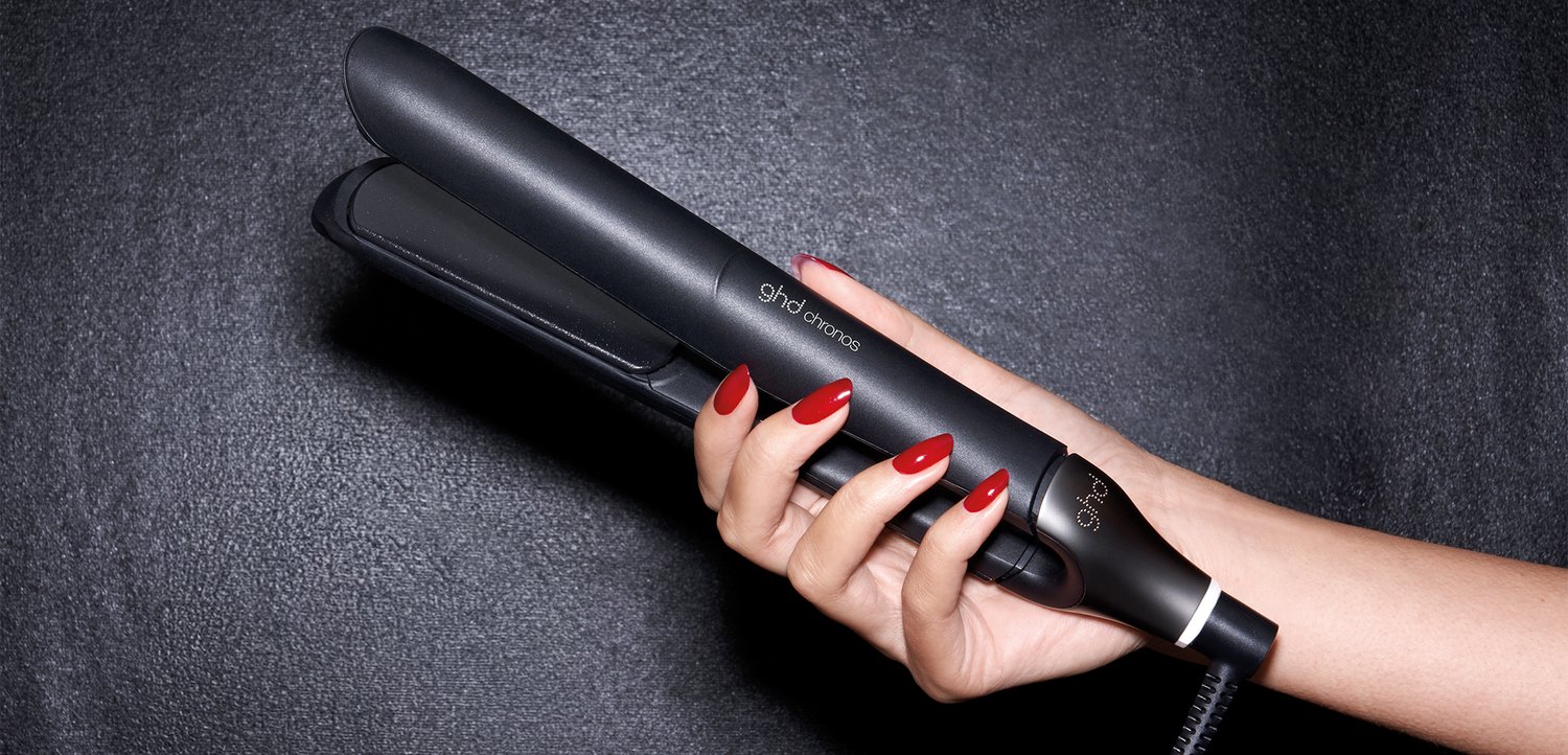 Main avec des ongles rouges tenant un fer à lisser ghd platinum+ noir sur un fond sombre.