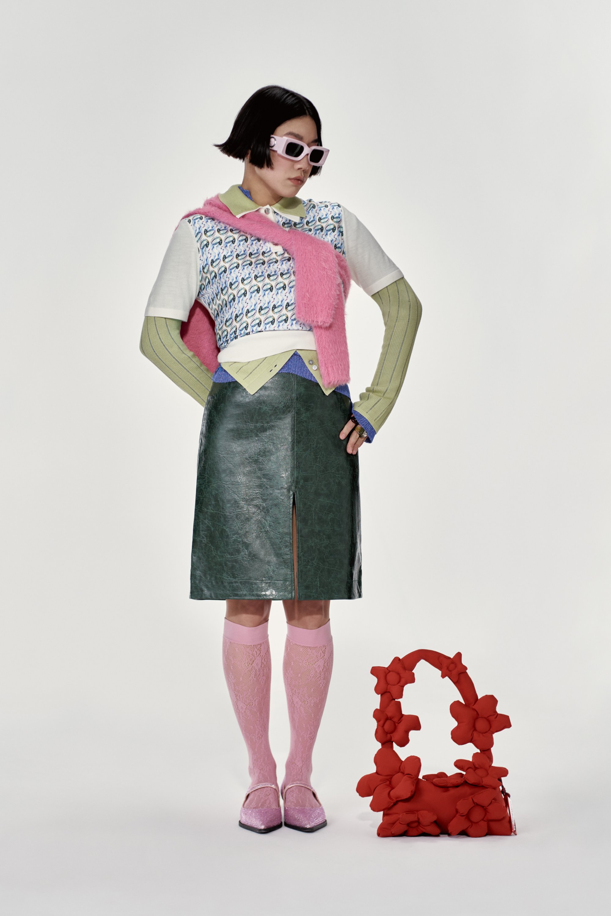 Conjunto colorido que incluye un top corto con patrón, falda corta de cuero sintético verde con una abertura, medias rosas y un bolso floral rojo.