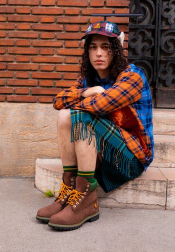 Ein junger Mann mit lockigem Haar sitzt auf Stufen, trägt ein kariertes Hemd, einen bunten Rock und braune Stiefel. Eine Ziegelwand dient als Hintergrund.