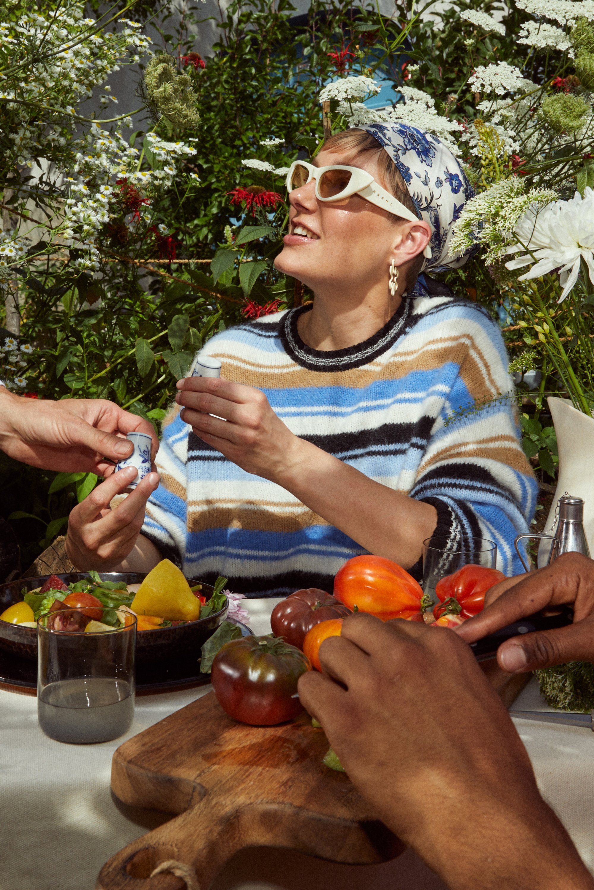 Una persona que lleva un suéter a rayas y gafas de sol sostiene un pequeño objeto, rodeada de coloridas verduras, flores y un vaso en una mesa.