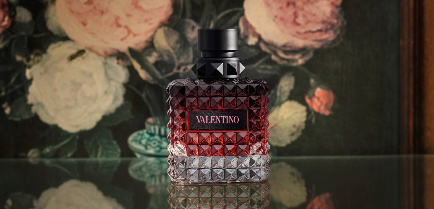 Glazen Valentino parfumflesje met een textuur van diamantpatroon, rode vloeistof binnenin, geplaatst op een reflecterend oppervlak met bloemrijke achtergrond.
