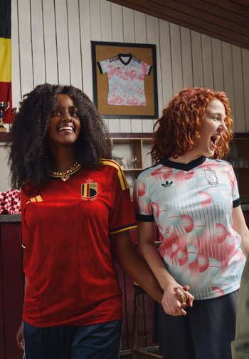 Twee vrouwen in voetbalshirts van België en Spanje houden elkaars hand vast en glimlachen in een informele binnenomgeving met een ingelijst shirt en trofeeën op de achtergrond.