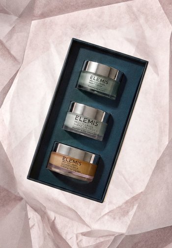 Tres frascos de cuidado de la piel Elemis Pro-Collagen en una caja oscura sobre papel tisú rosa arrugado.