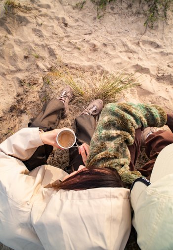 Een persoon zit op de zandgrond, met een mok koffie in de hand, gekleed in een beige jack en omringd door gras en zacht terrein.