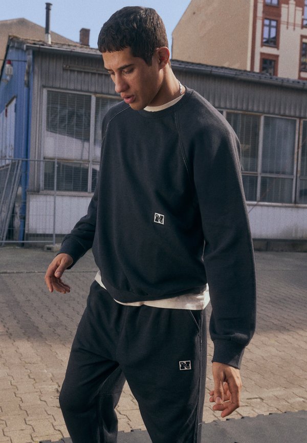 Un jeune homme en sweat-shirt gris et en survêtement marche dans une zone extérieure, les yeux baissés pensivement. Des bâtiments industriels se trouvent en arrière-plan.