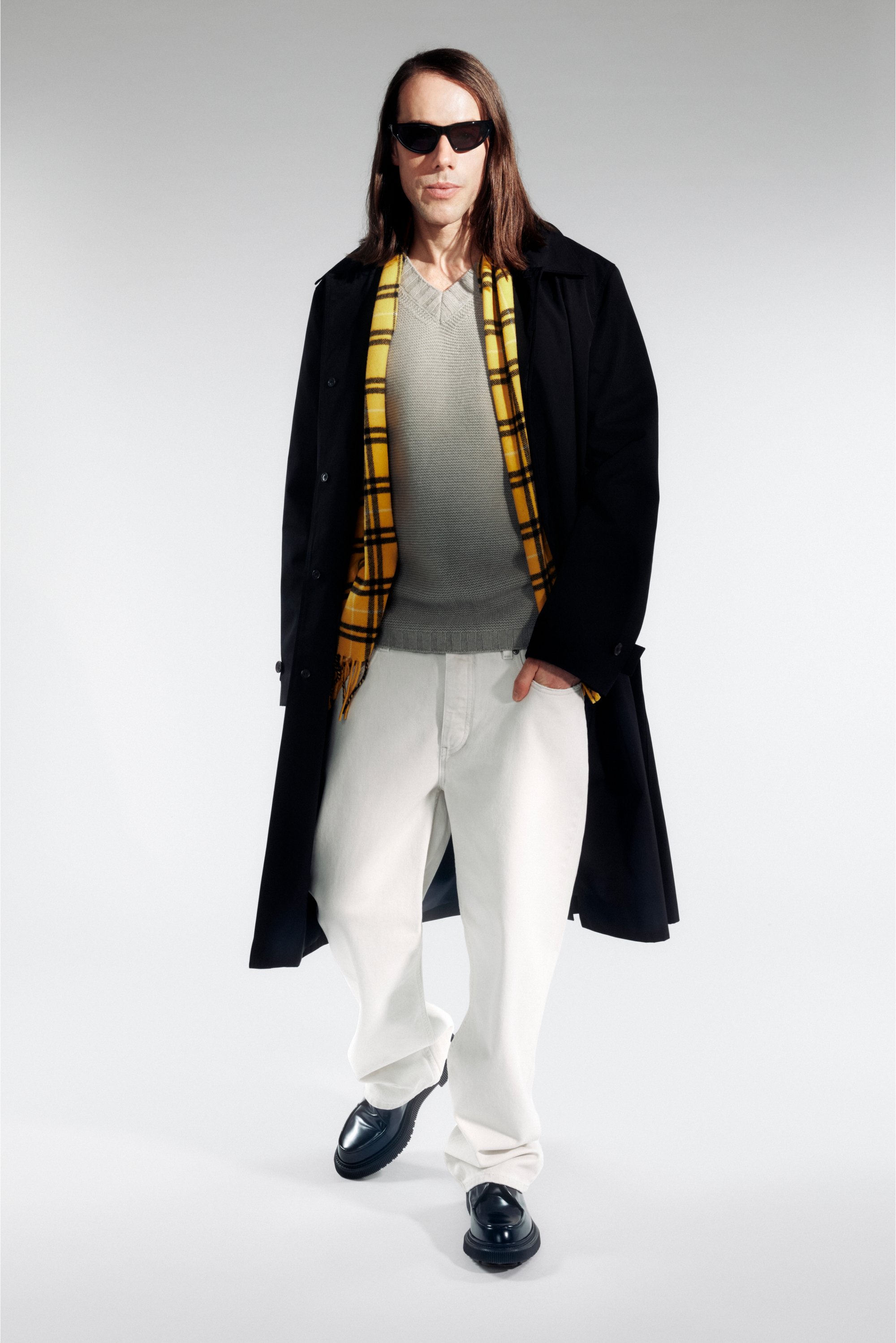 Una persona con cabello largo lleva gafas de sol, un abrigo negro, un suéter gris y una bufanda de cuadros amarillos, combinados con pantalones blancos y zapatos negros.
