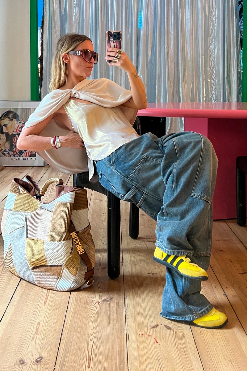 Mujer con gafas de sol y top de satén reclinada en una silla negra junto a un bolso de patchwork, una mesa colorida y una obra de arte enmarcada sobre el suelo de madera.
