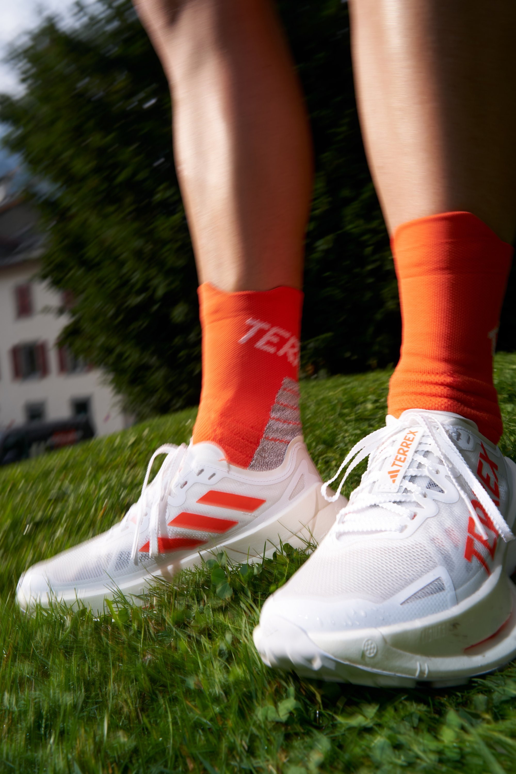 White athletic shoes with orange accents and three stripes, paired with high orange socks featuring a grey pattern, placed on green grass.