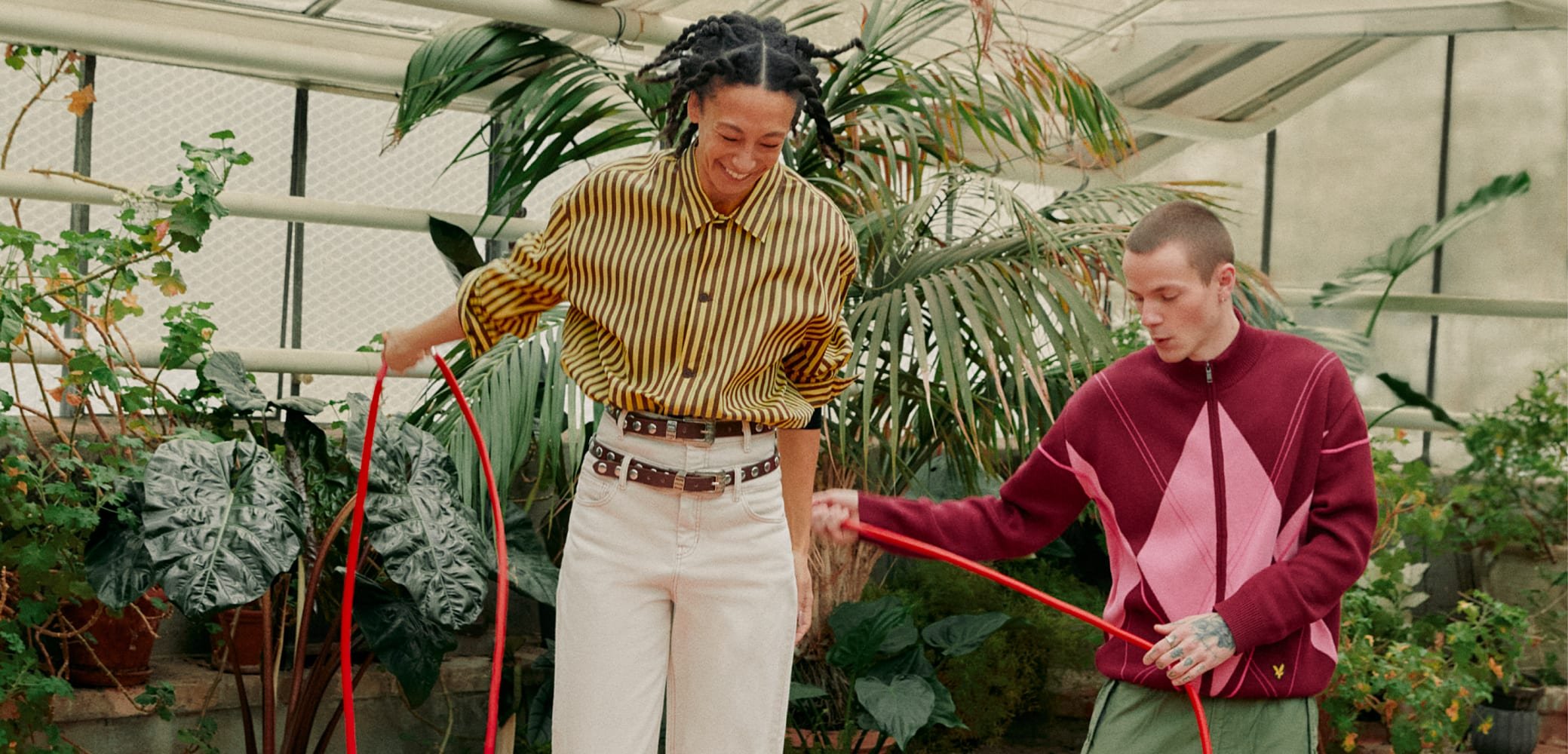 Zwei Personen lächeln und halten einen roten Gartenschlauch in einem Gewächshaus, umgeben von großen grünen Pflanzen.