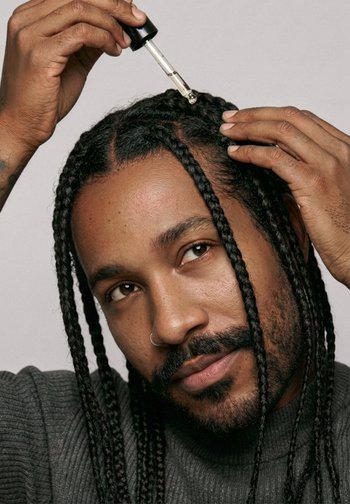 Uomo con capelli intrecciati applica un liquido da un contagocce direttamente sul cuoio capelluto, tenendo i capelli da parte con una mano.